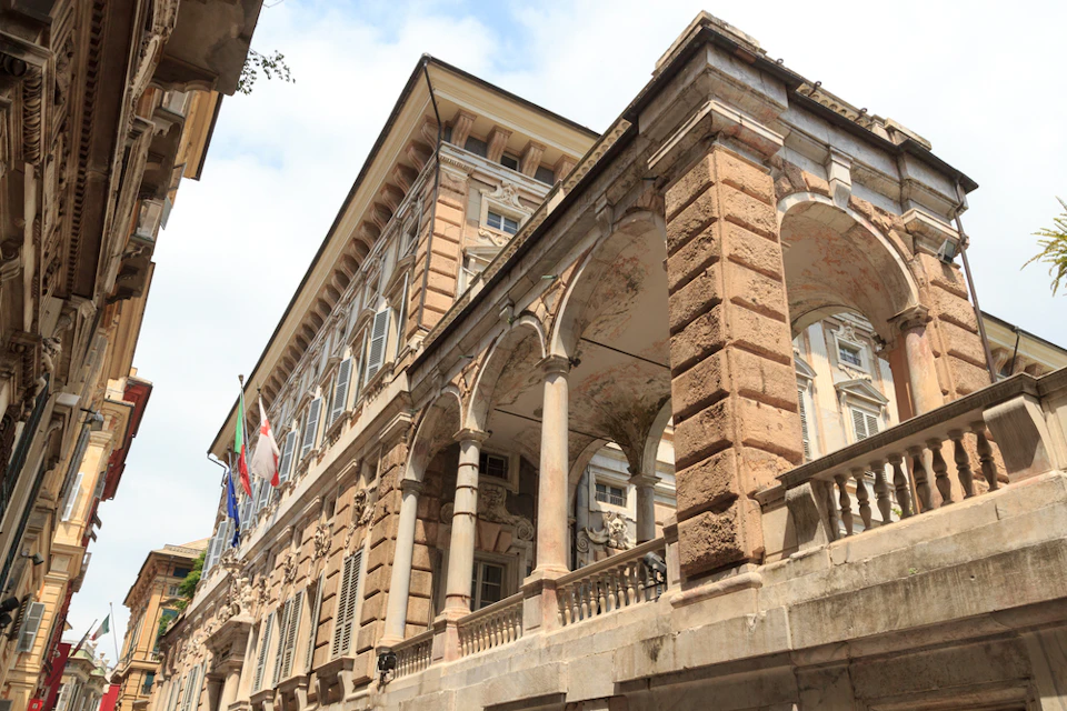 Elegant historic building with arched columns and flags, featuring ornate architectural details and windows. Blue sky in the background.