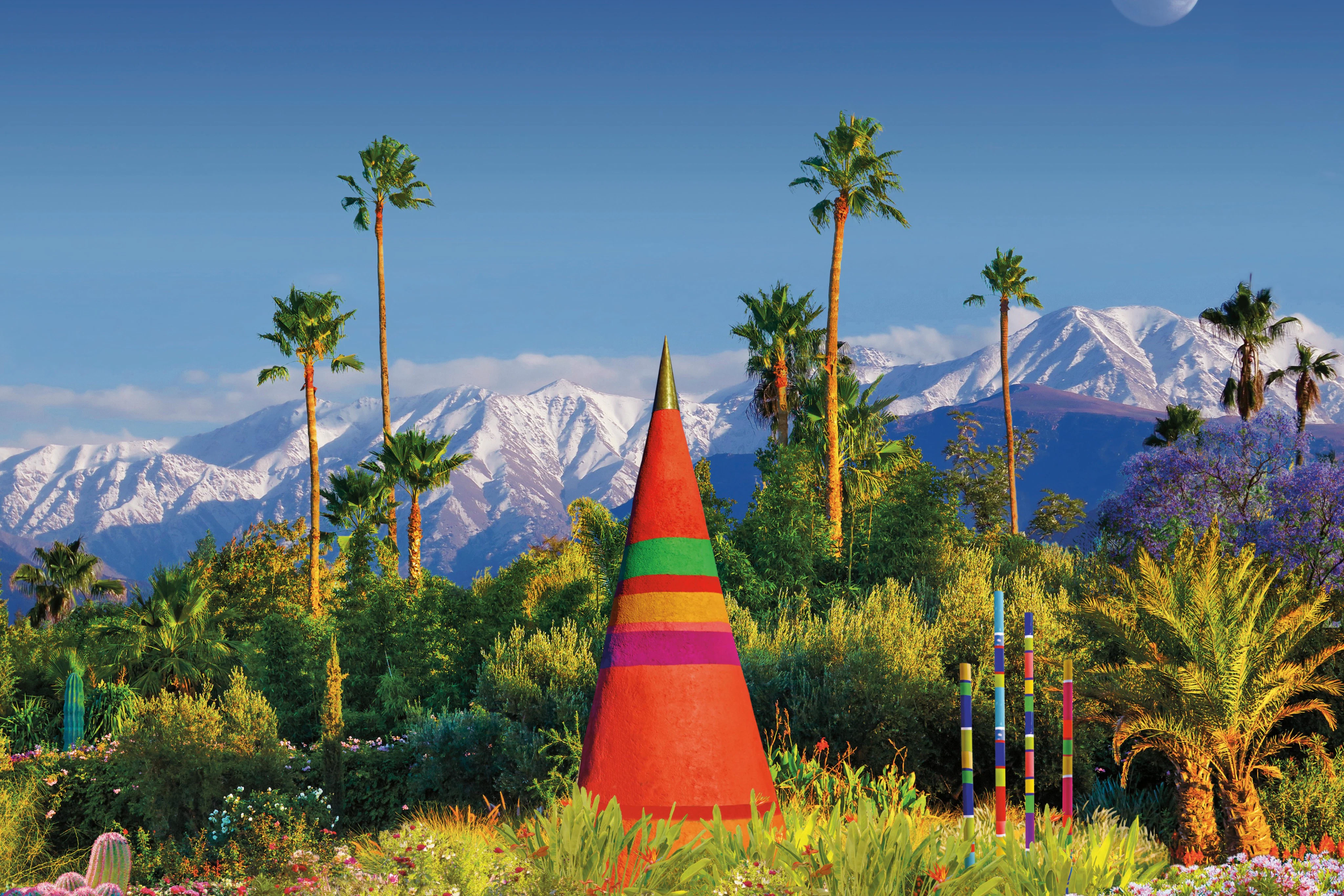 A colorful cone-shaped sculpture surrounded by greenery and palm trees, with snow-capped mountains in the background.