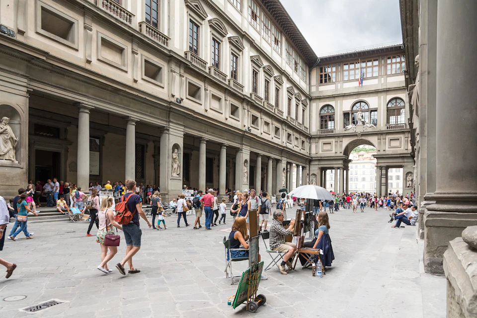 Crowded courtyard with people walking and seated, surrounded by columns and historic buildings; street artists painting in the foreground.
