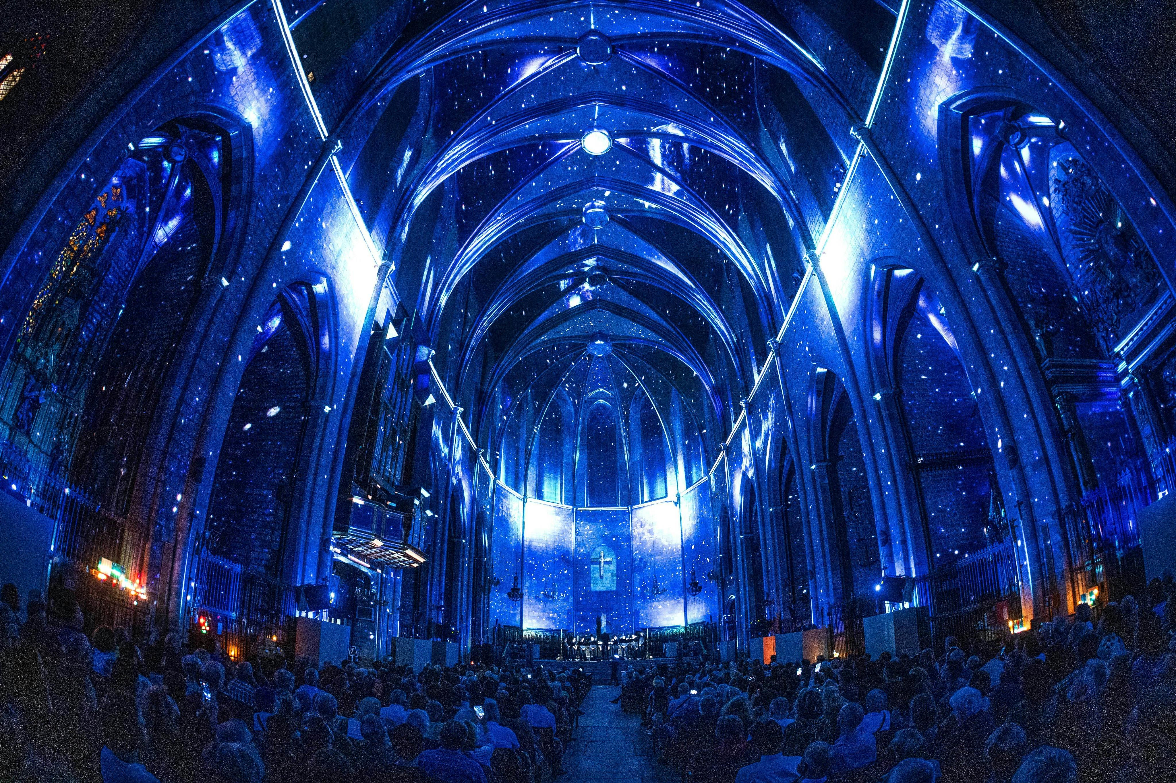 L'intérieur d'une cathédrale baignée d'une lumière bleue avec un effet de ciel étoilé sur les murs et le plafond, remplie de spectateurs assis.