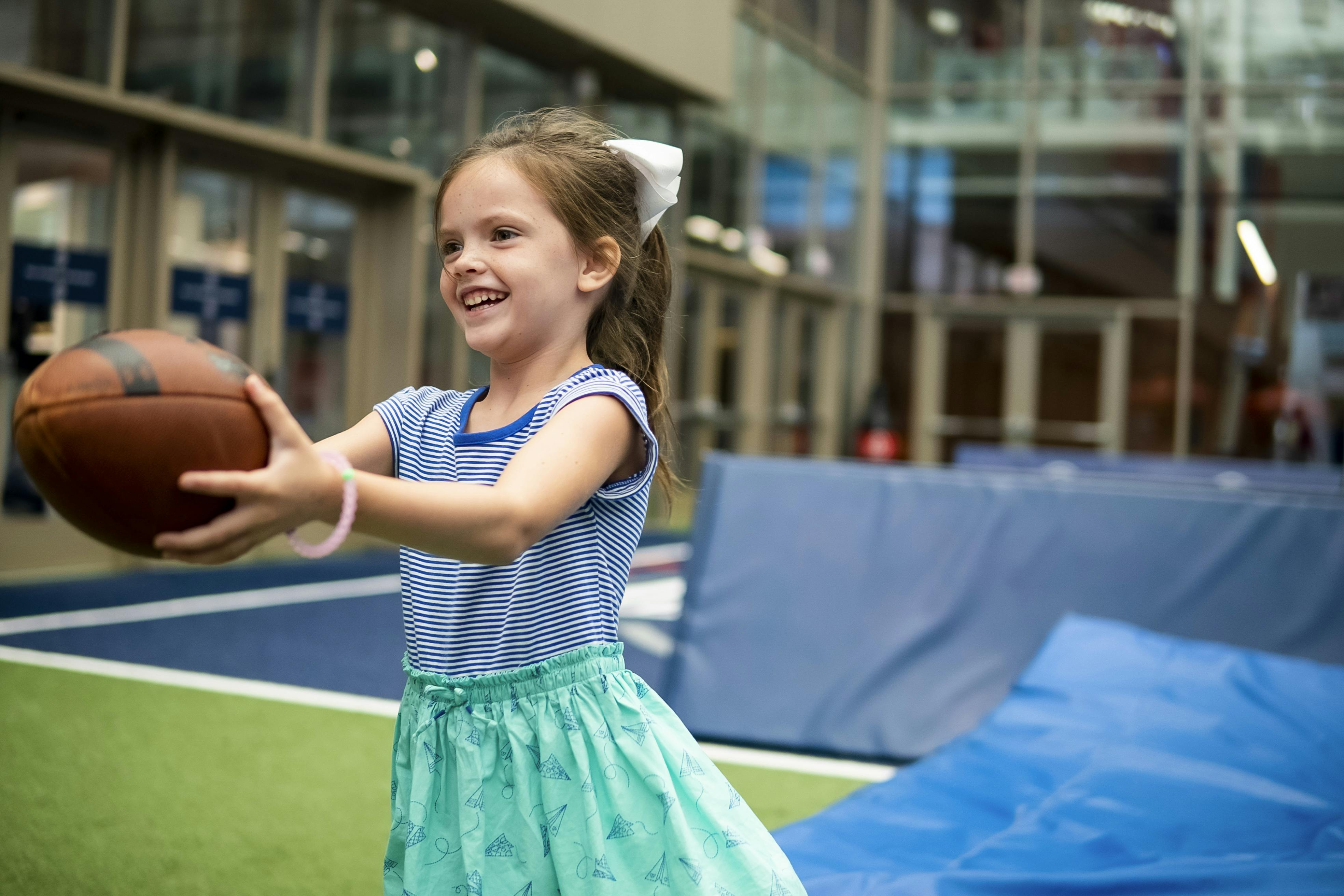Mladá dívka s bílou mašlí ve vlasech se usmívá, zatímco drží fotbalový míč na krytém trávníkovém hřišti.