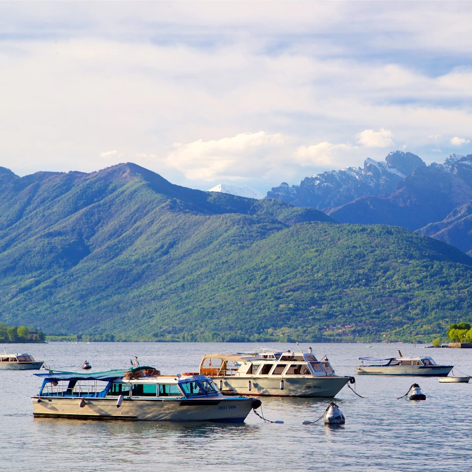 Multiple boats anchored on a calm lake with green, forest-covered mountains and a partly cloudy sky in the background.