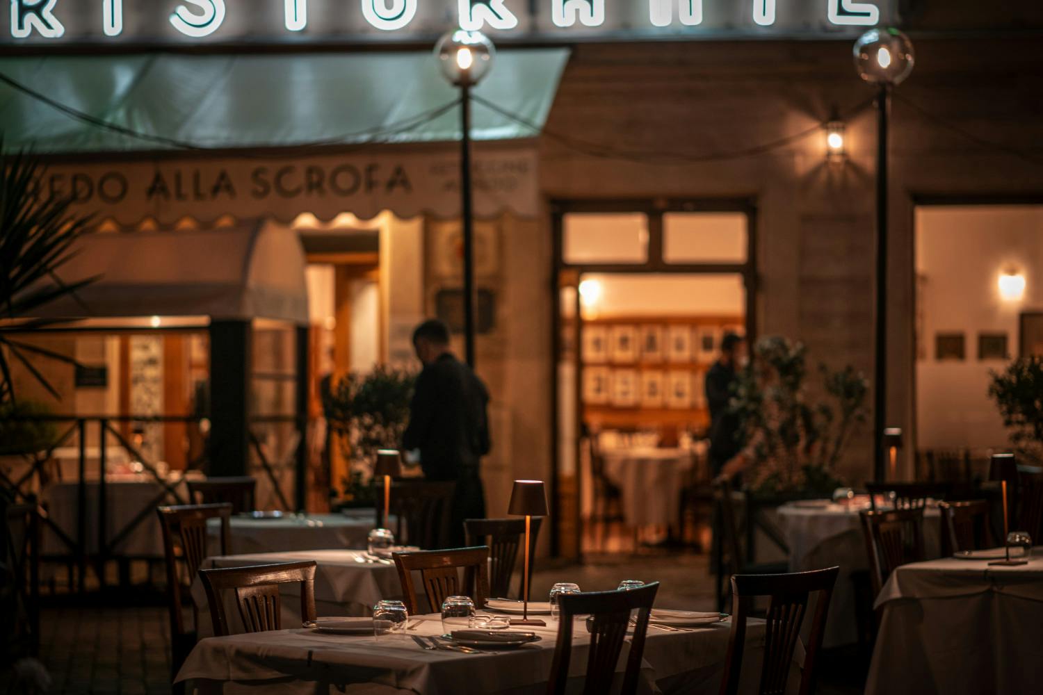 Тускло освещенный ресторан на открытом воздухе с установленными столами и стеклянной посудой, двумя людьми на заднем плане и частично видимой подсвеченной вывеской выше.