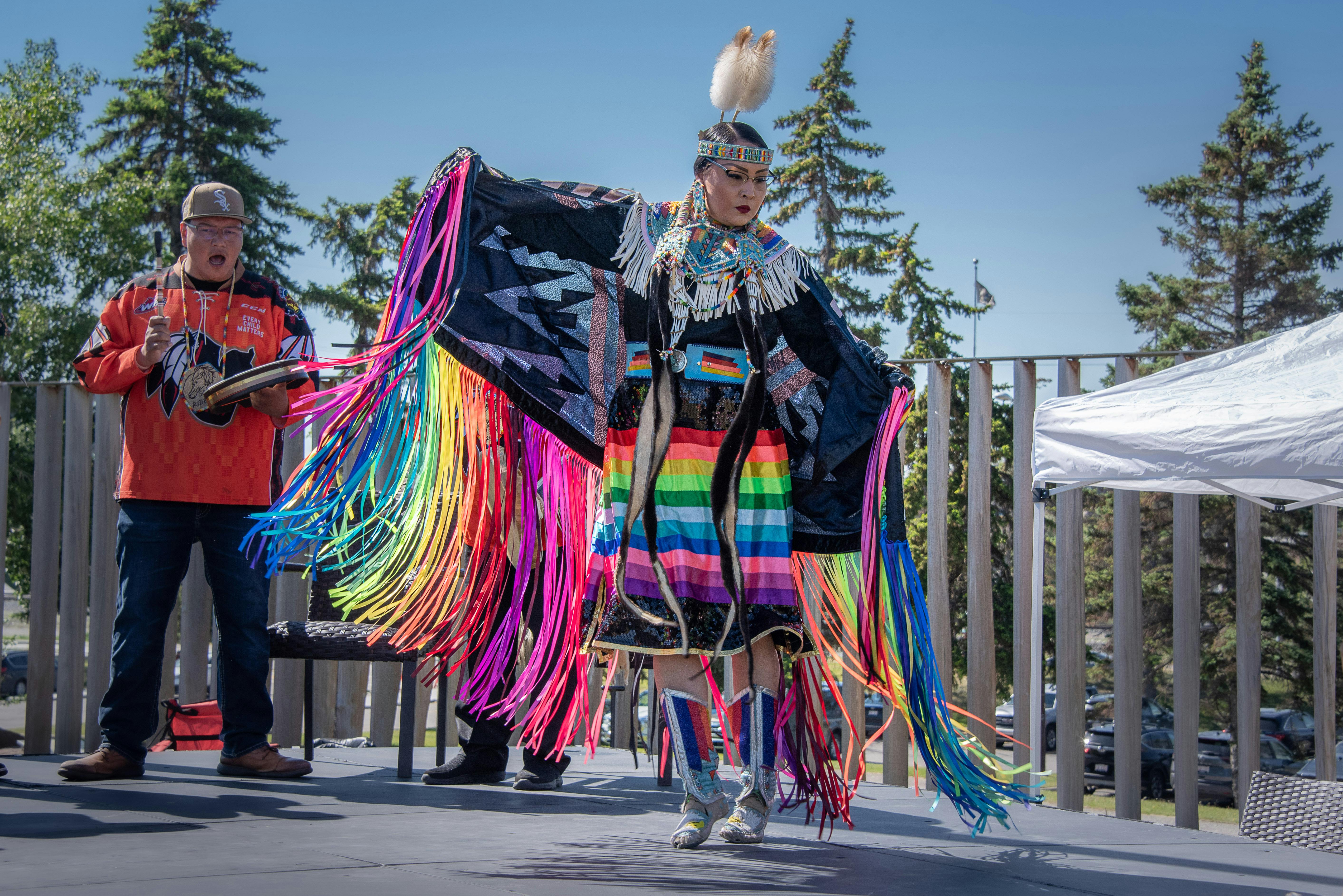 A person dances wearing a vibrant, multicolored outfit with long fringes on a stage, set outdoors against a backdrop of trees.