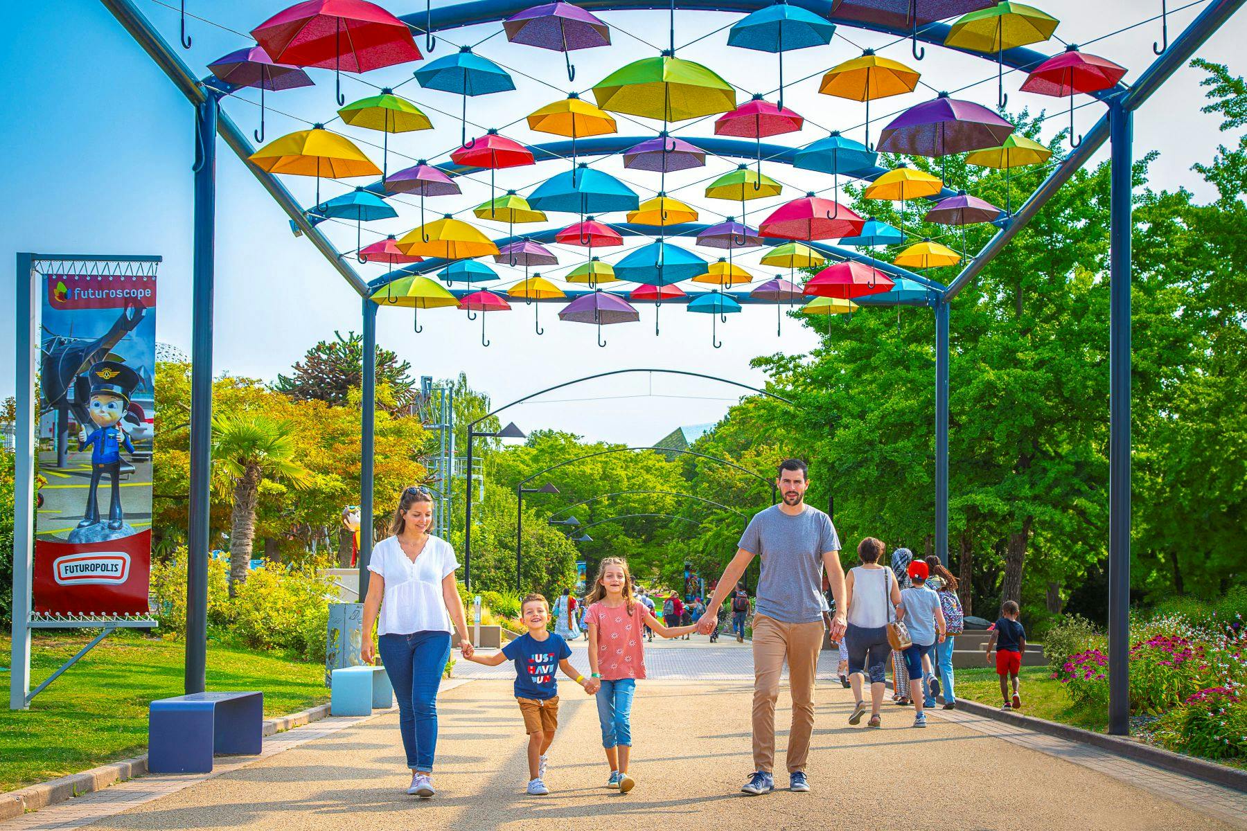Family walking under colorful hanging umbrellas in a park with greenery and other people in the background.