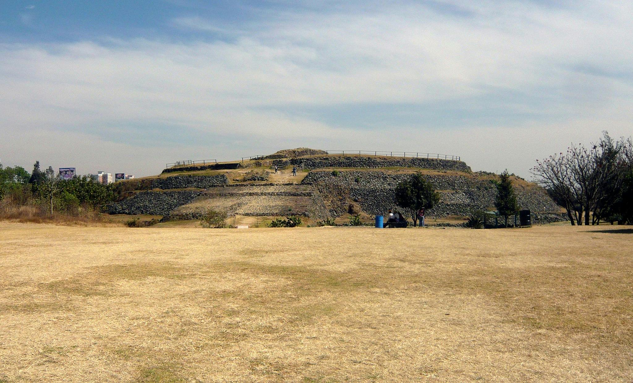 Stone pyramid with people walking, surrounded by dry grass and sparse trees under a partly cloudy sky.