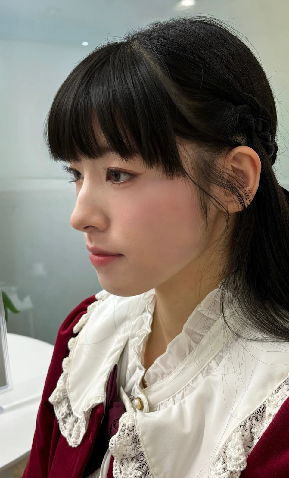 A profile view of a person with dark hair and bangs, wearing a white blouse with red details, in a softly lit indoor setting.
