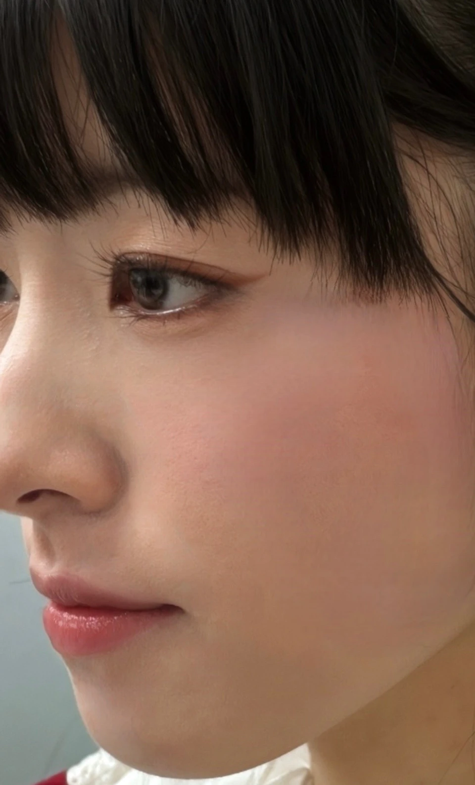A profile view of a person with dark hair and bangs, wearing a white blouse with red details, in a softly lit indoor setting.