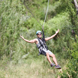 A person wearing a helmet and harness ziplining through a lush green forest, smiling with arms outstretched.