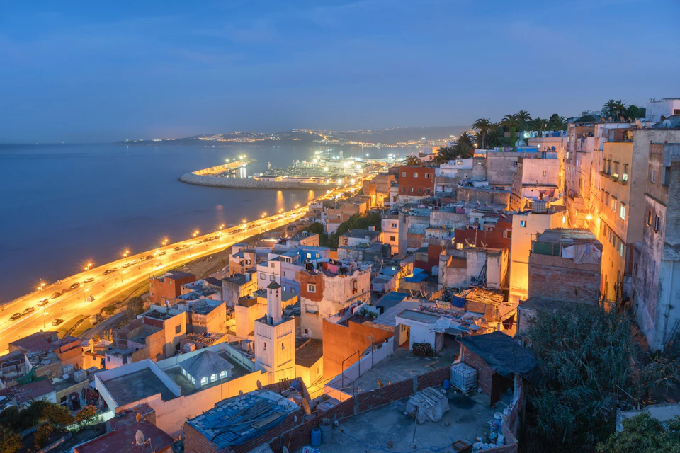 A coastal city at dusk with illuminated buildings and a lit-up road along the shoreline, featuring vibrant houses and a calm sea.