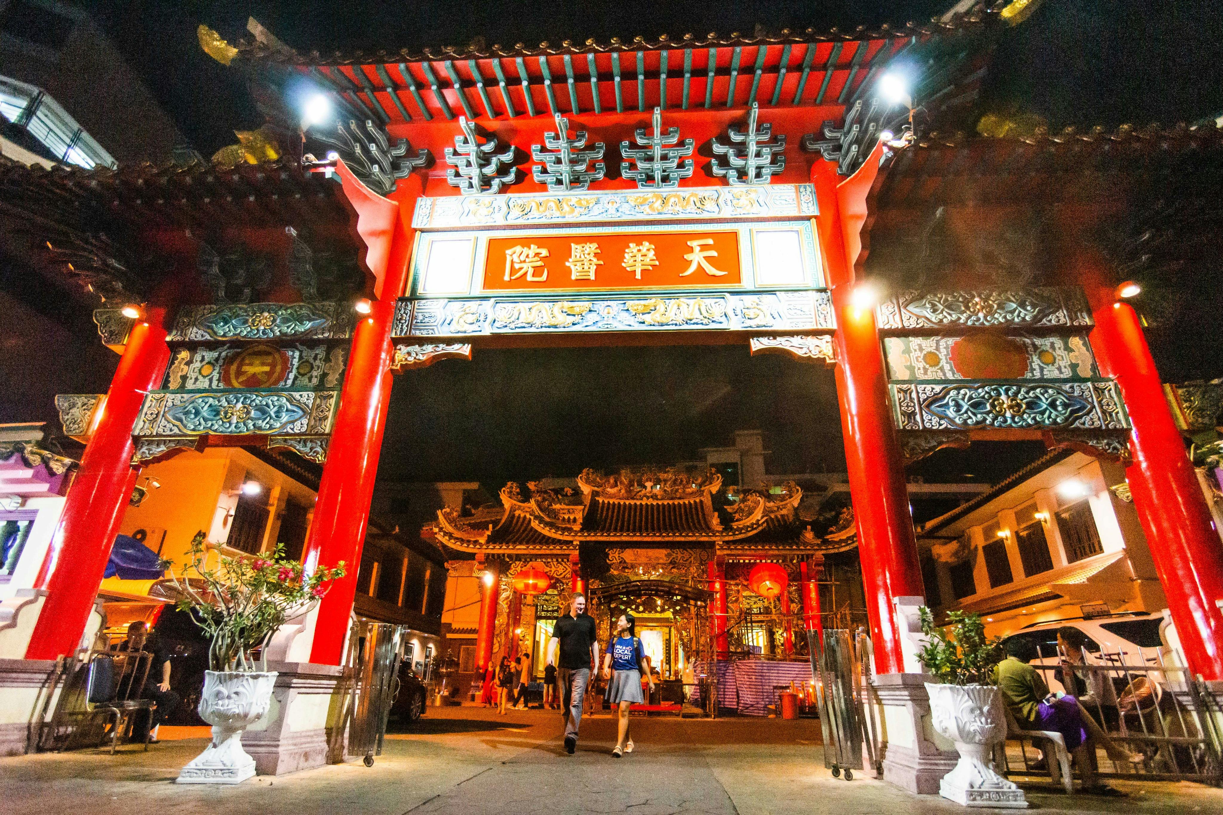 晚上，两个人走过一个明亮的红色和金色的寺庙大门，上面有复杂的屋顶细节和汉字。