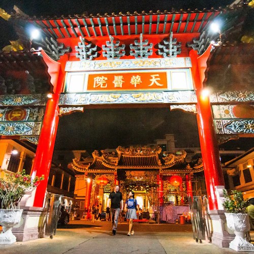 Zwei Personen gehen nachts durch ein hell erleuchtetes rot-goldenes Tempeltor mit verschlungenen Dachdetails und chinesischen Schriftzeichen.