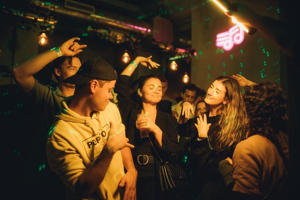 People dance in a dimly lit club with neon lights and glowing wristbands. A neon music note is visible on the wall.