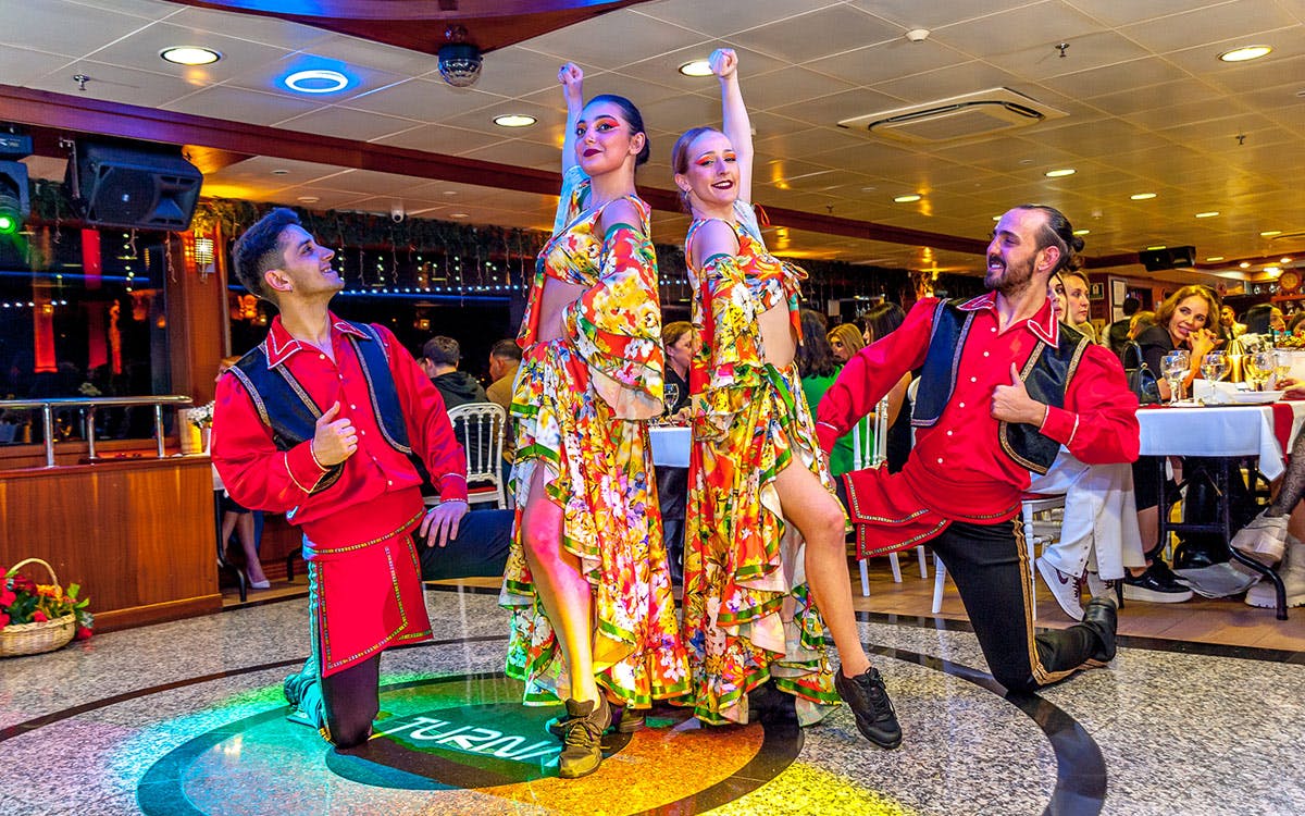 Four dancers in colorful floral costumes and red shirts perform on a lit dance floor in a brightly decorated indoor space.