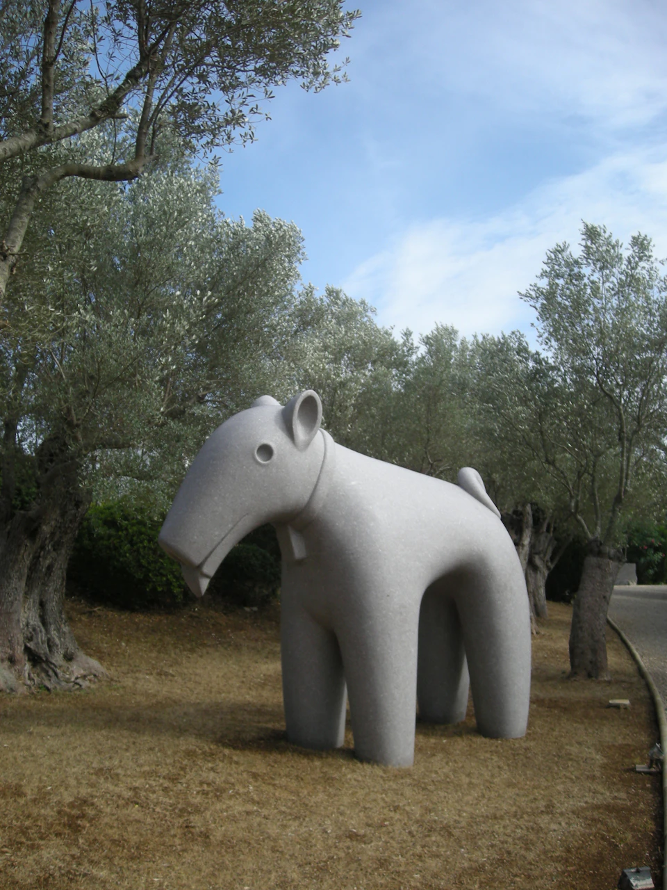 Stone sculpture of a stylized animal surrounded by olive trees on a dry, grassy area under a partly cloudy sky.