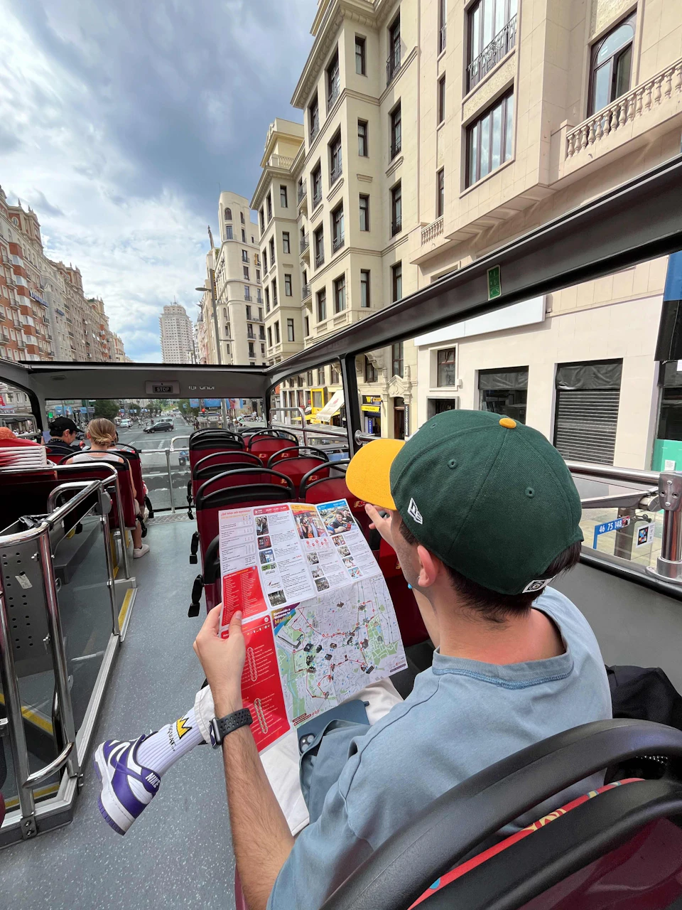 A person wearing a green and yellow cap reads a map while sitting on the upper deck of an open-top tour bus in a city.