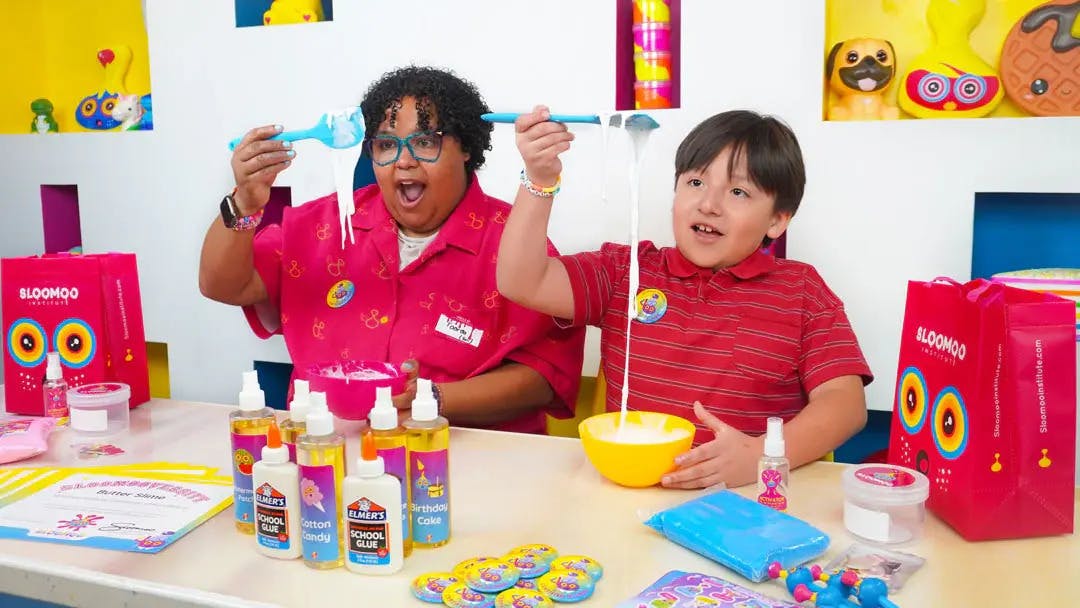 Dva lidé vyrábějící sliz u stolu s lepidlem, miskami a barevnými pomůckami. Člověk vypadá nadšeně, když drží pružný sliz.