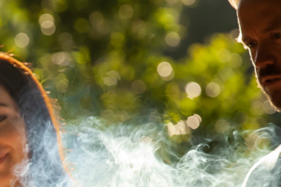A woman and man standing outdoors, observing smoke rising from a burning object held by an unseen person.