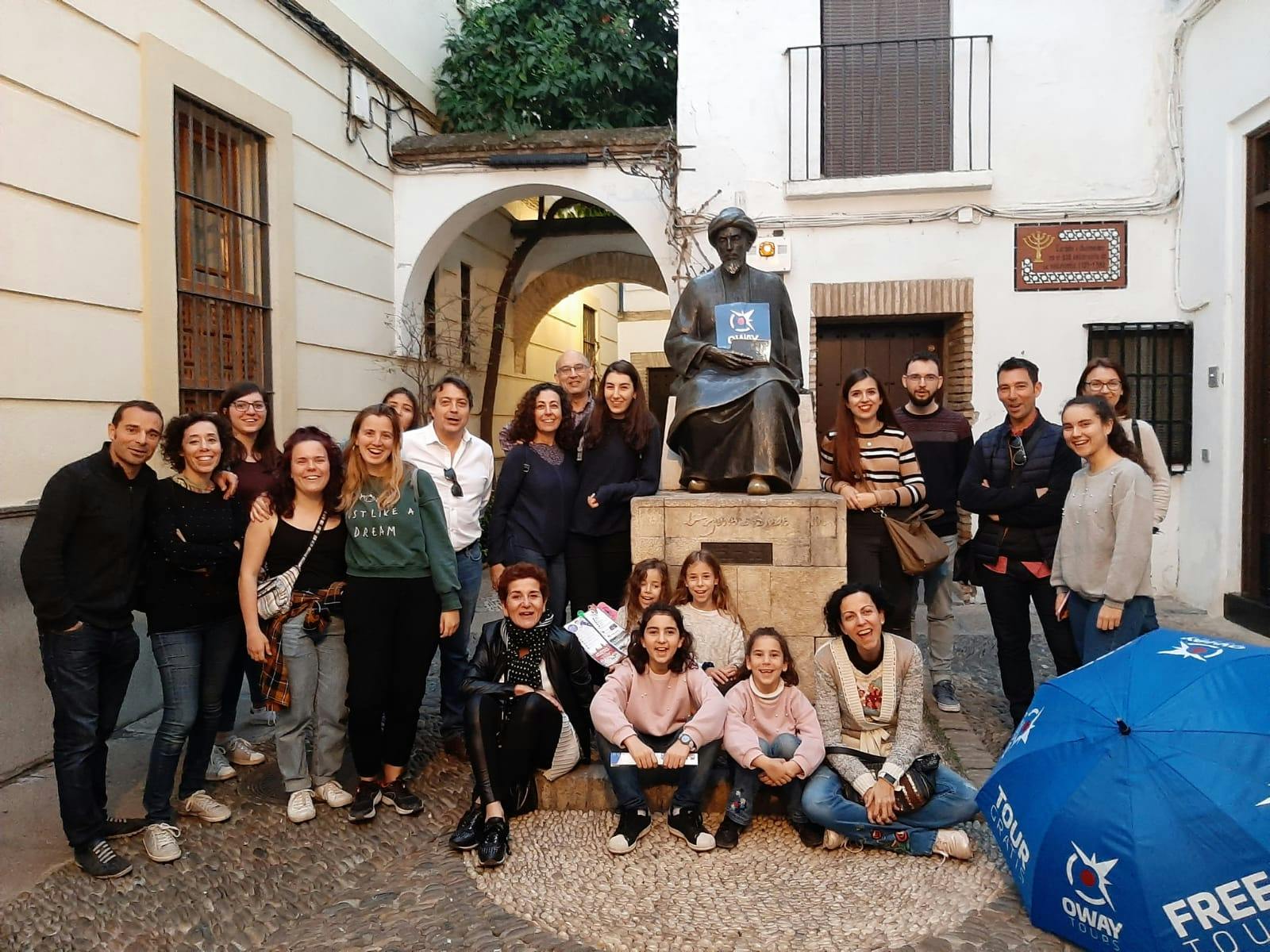 Group of people posing around a seated statue in a narrow street with white buildings and an archway in the background.