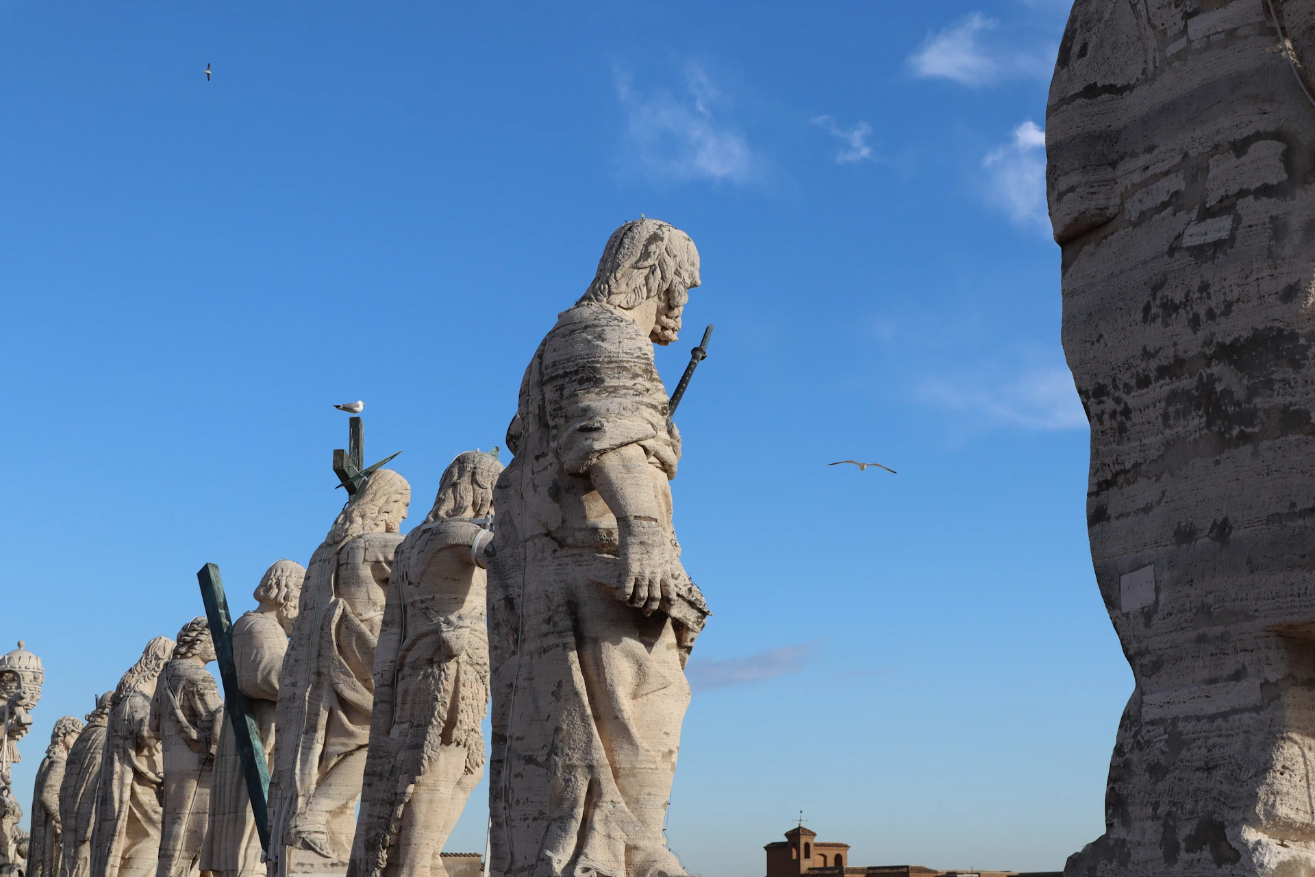 A row of large stone statues against a blue sky, with a bird flying nearby and a distant building visible on the horizon.