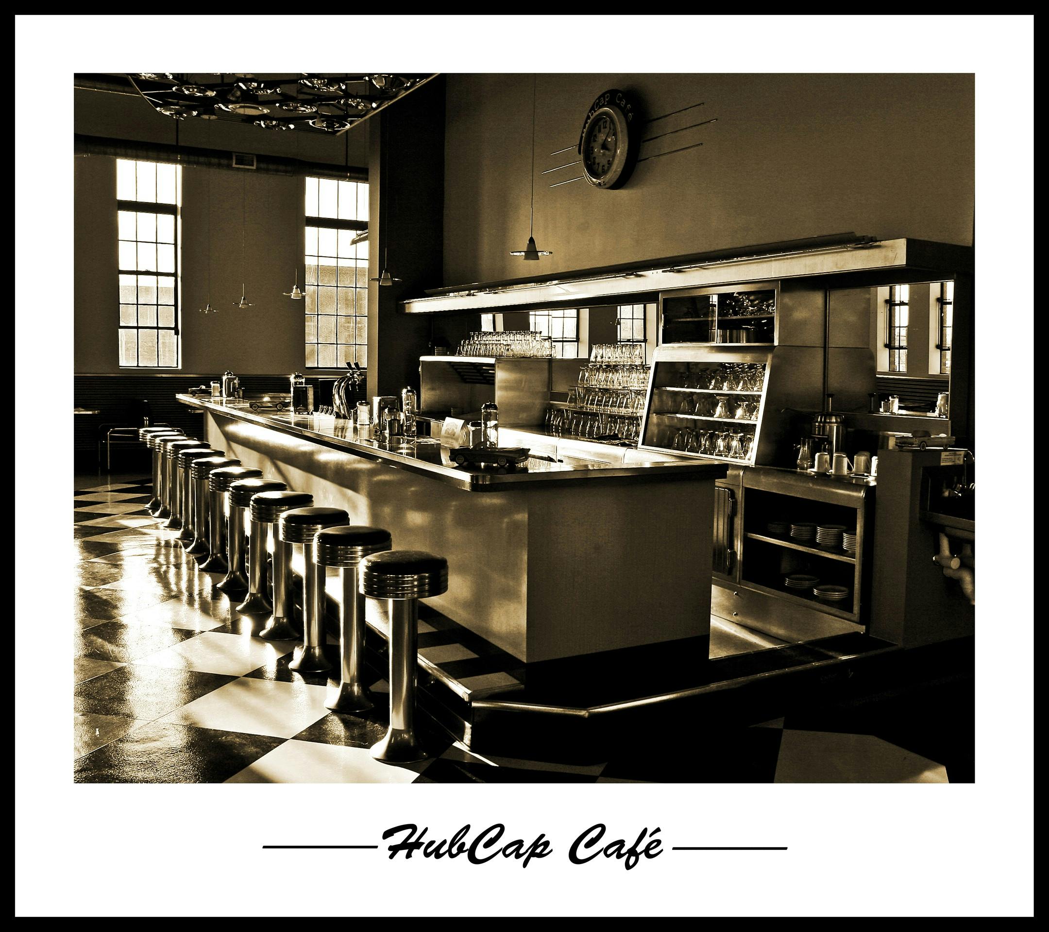 Μια ρετρό καφετέρια με μακρύ πάγκο, χρωμιωμένα σκαμπό μπαρ, καρό πάτωμα και μεγάλα παράθυρα. Το "HubCap Café" έχει λεζάντα παρακάτω.