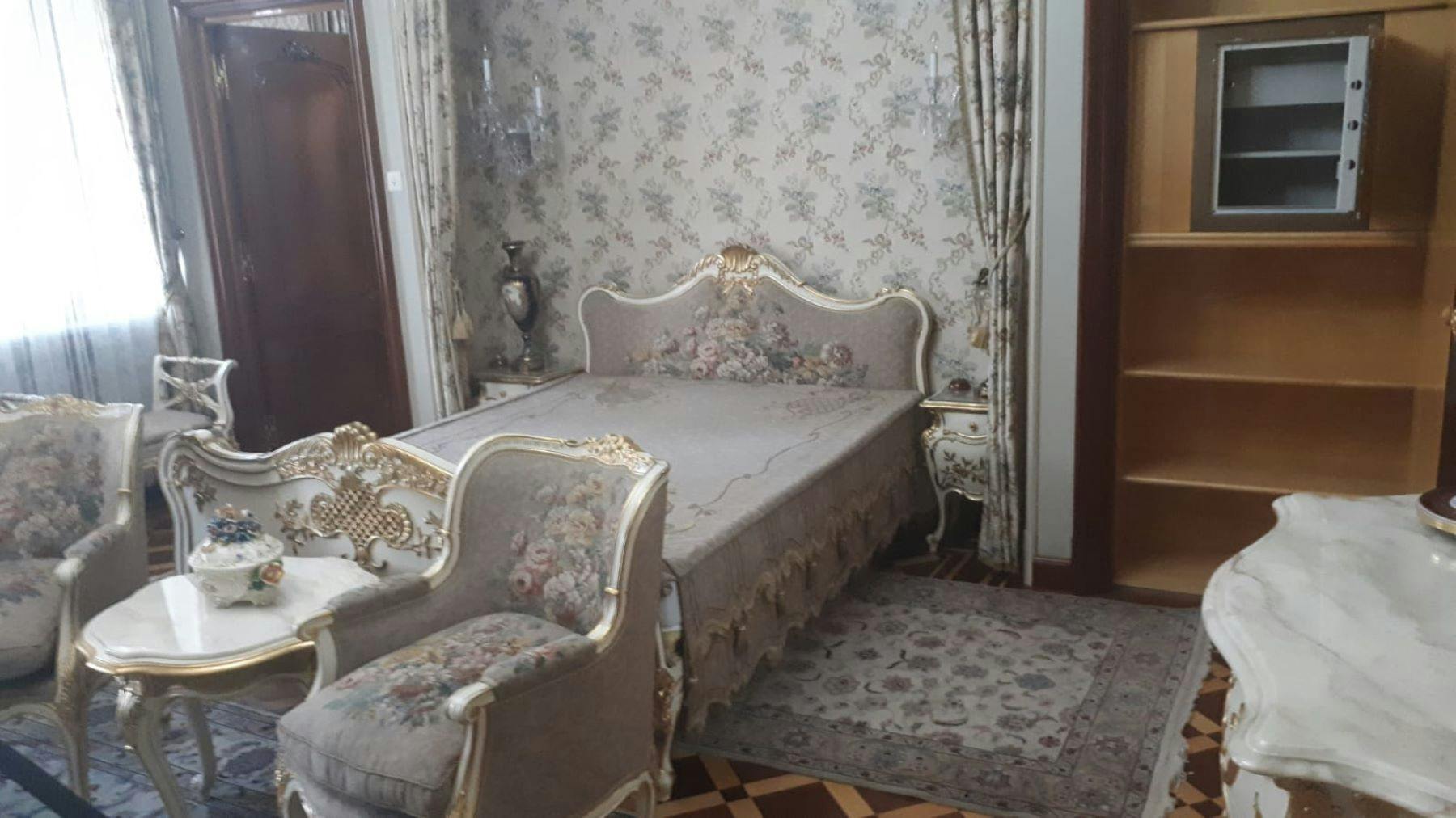 Dormitori adornat amb paper pintat floral, un llit tallat amb tapisseria floral, butaques a joc, taules auxiliars i una catifa estampada.