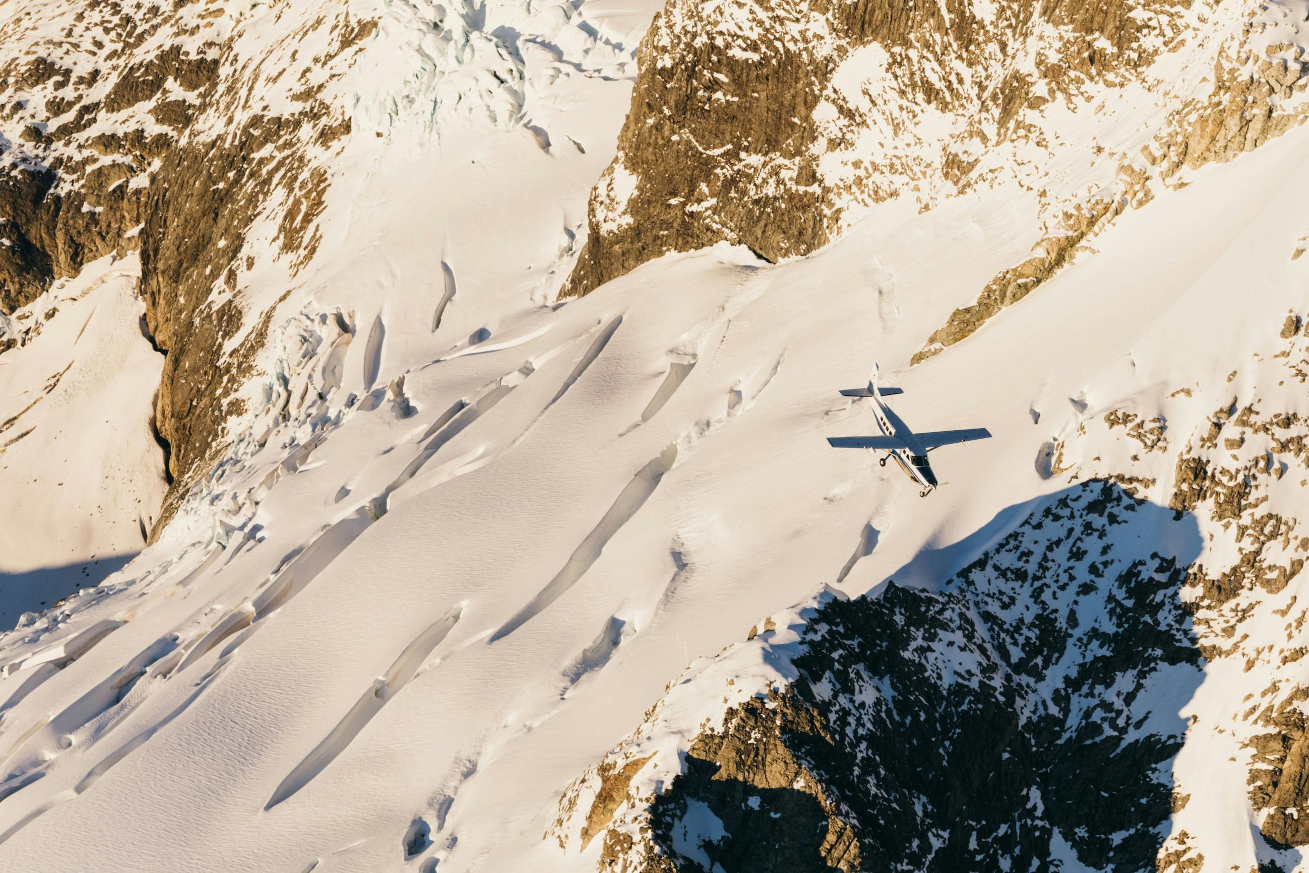 A small aircraft flying over a snow-covered mountain range with visible crevices and rocky peaks in sunlight.