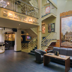 Restauranginteriör med moderna sittplatser, stor väggmålning, spiraltrappa och "TJÄNA ALLA" -skylt. Inramade konstverk finns på väggarna.