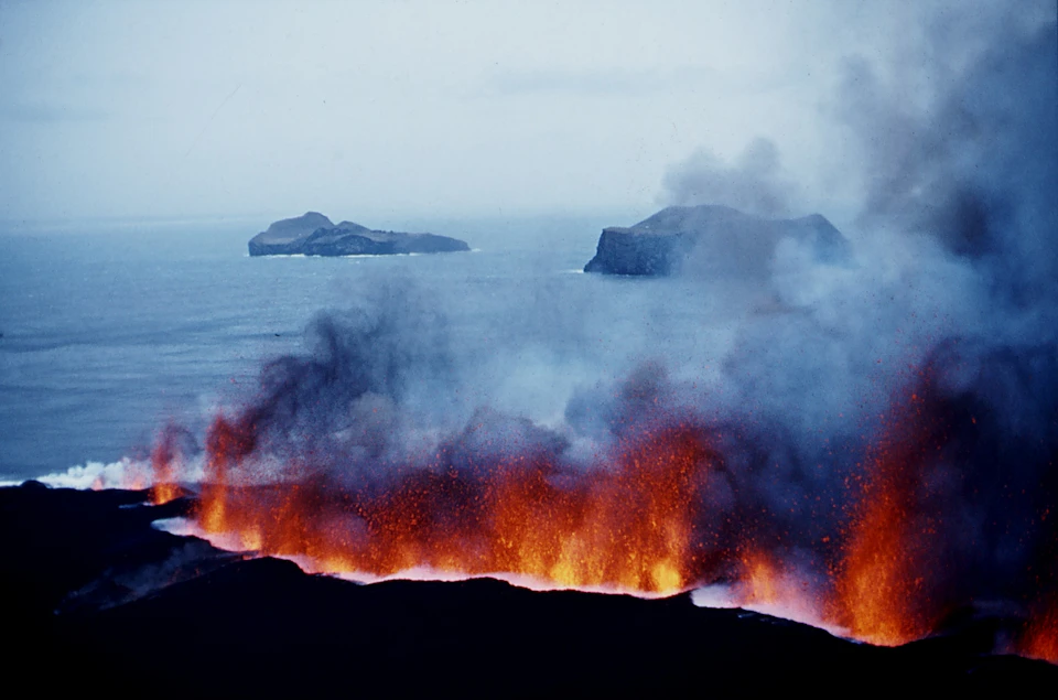 Erupting volcano spewing lava and smoke near a coast with two distant islands in view against a cloudy sky.