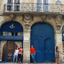Fyra personer står utanför den utsmyckade blå ingången till "Les Caves du Louvre" i Paris, bredvid en stor trädörr.