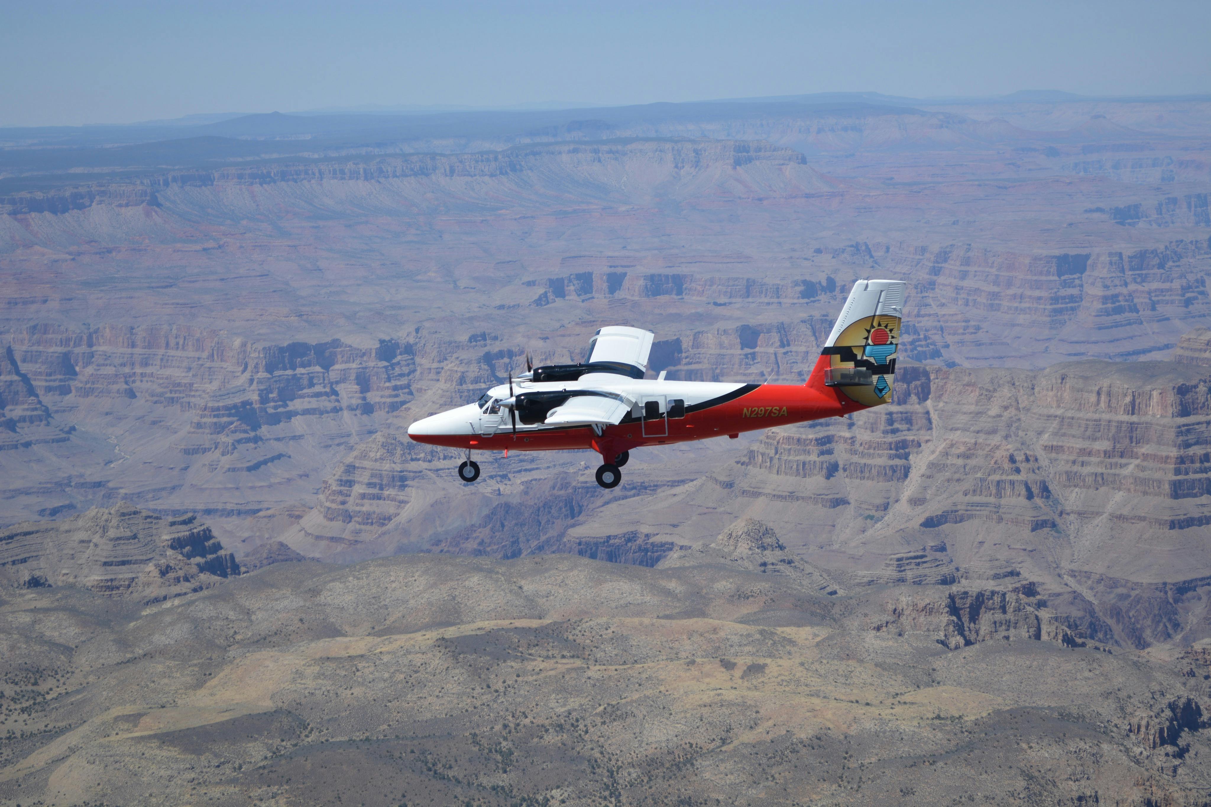 A small white and red airplane flies over the expansive Grand Canyon landscape.