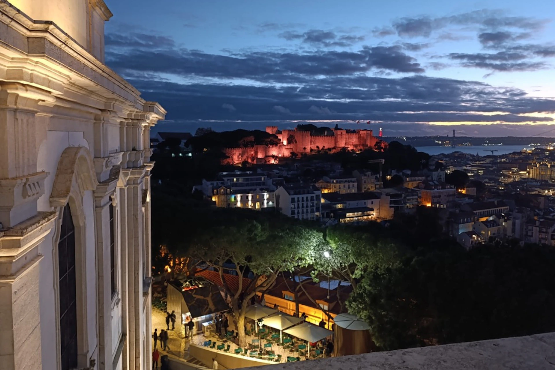A cityscape at dusk with a lit hillside castle, buildings, trees, and a partially visible stone structure on the left.