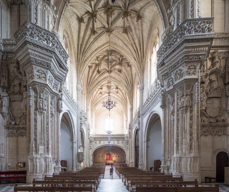 A person walks toward the altar in a grand, ornately decorated cathedral with vaulted ceilings, detailed carvings, and wooden pews.