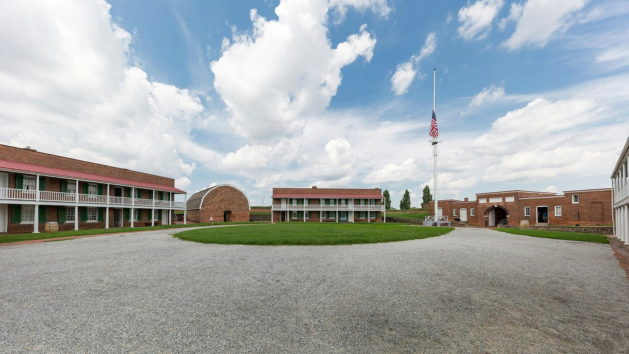 Une cour de fort historique avec des bâtiments en briques, un mât avec un drapeau américain et un ciel clair avec des nuages épars.