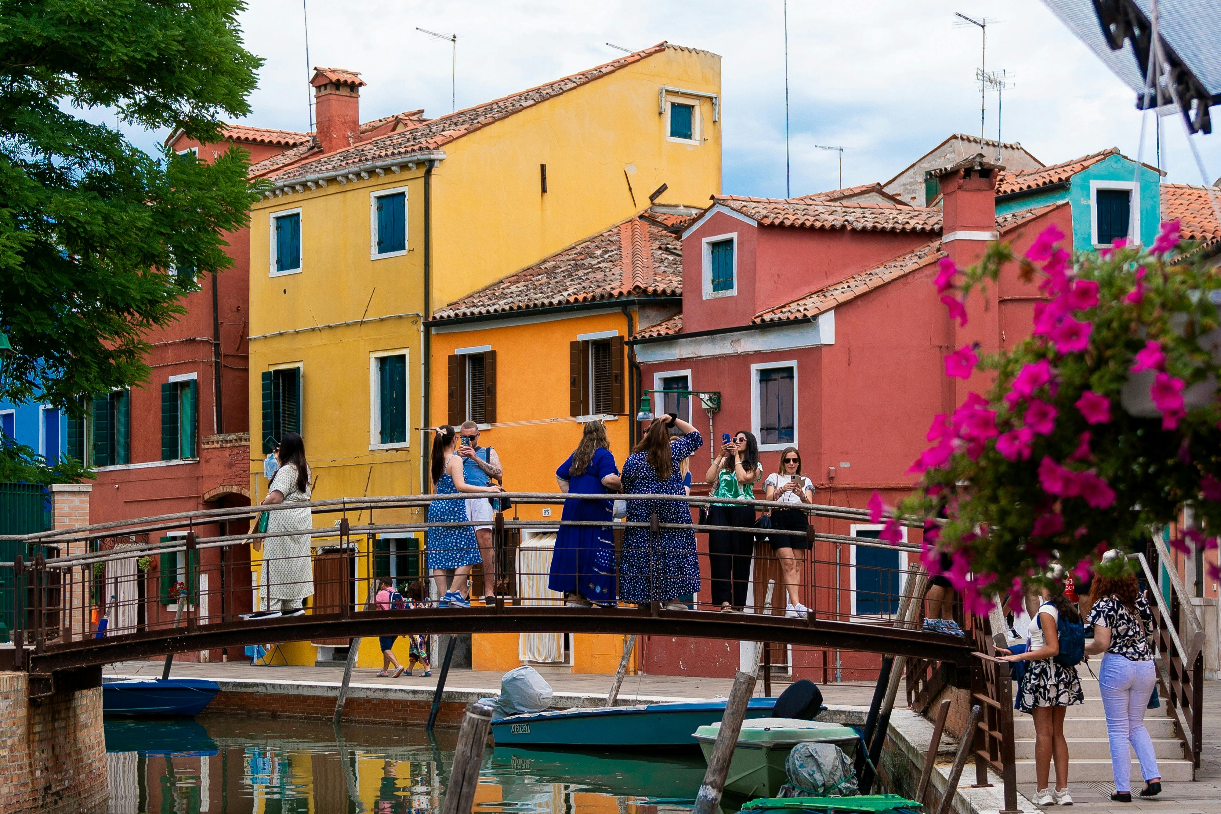 People are gathered on a bridge in front of brightly colored houses, taking photos and conversing. Flowers and boats are nearby.