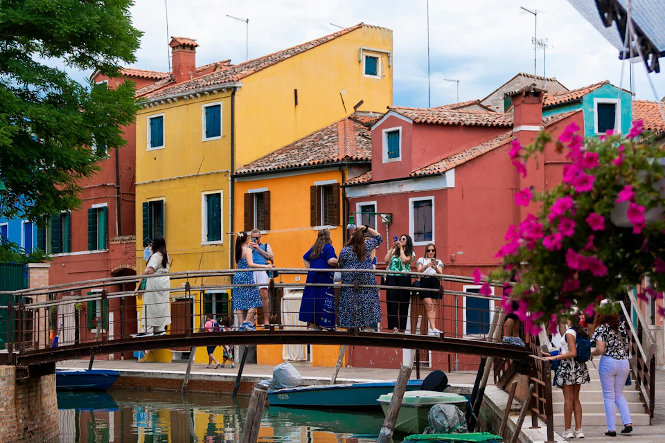 People are gathered on a bridge in front of brightly colored houses, taking photos and conversing. Flowers and boats are nearby.