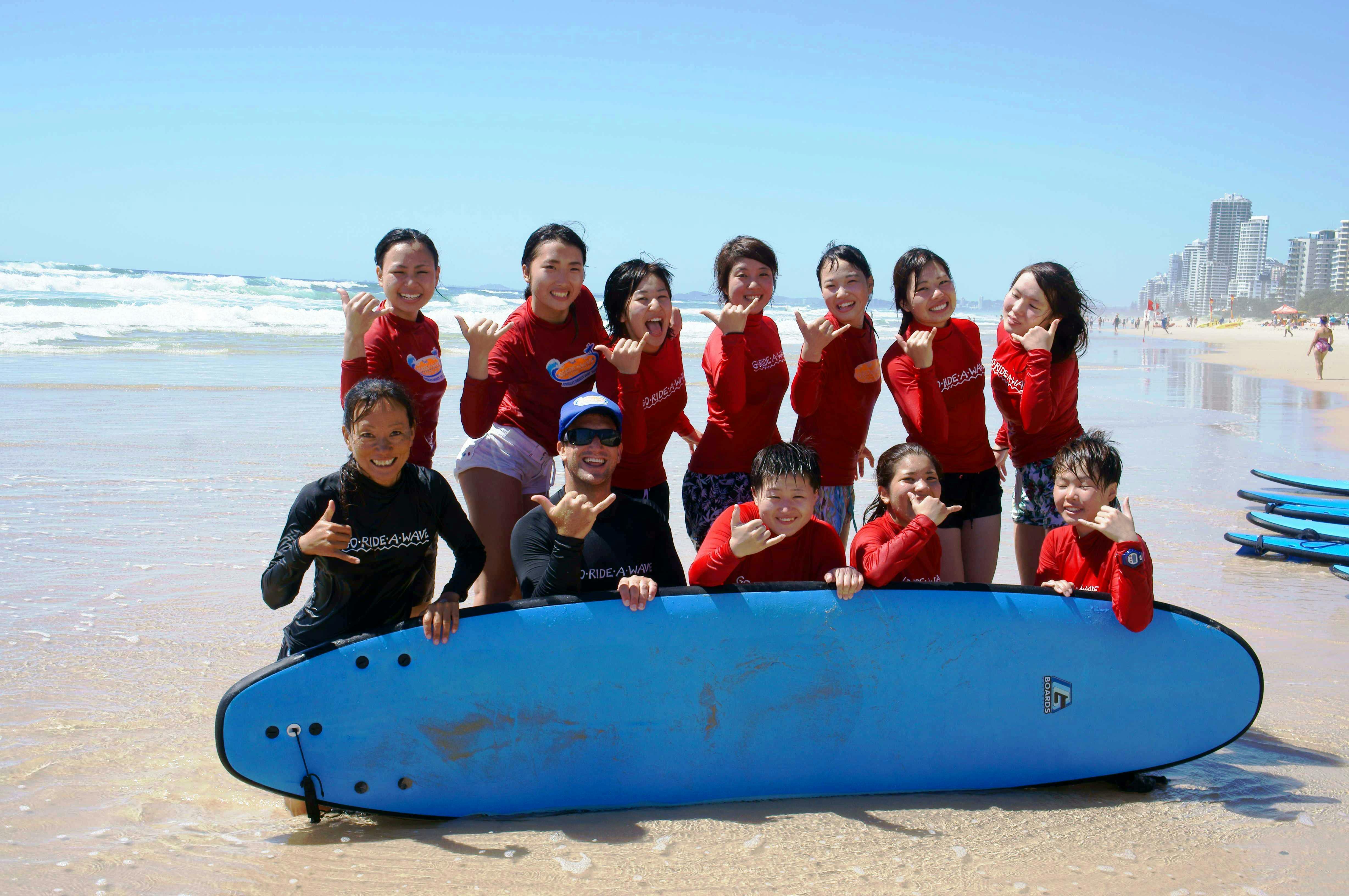 一群穿着红色衬衫的人在海滩上摆着冲浪板，做着沙卡招牌，微笑着。 波浪在背景中可见。