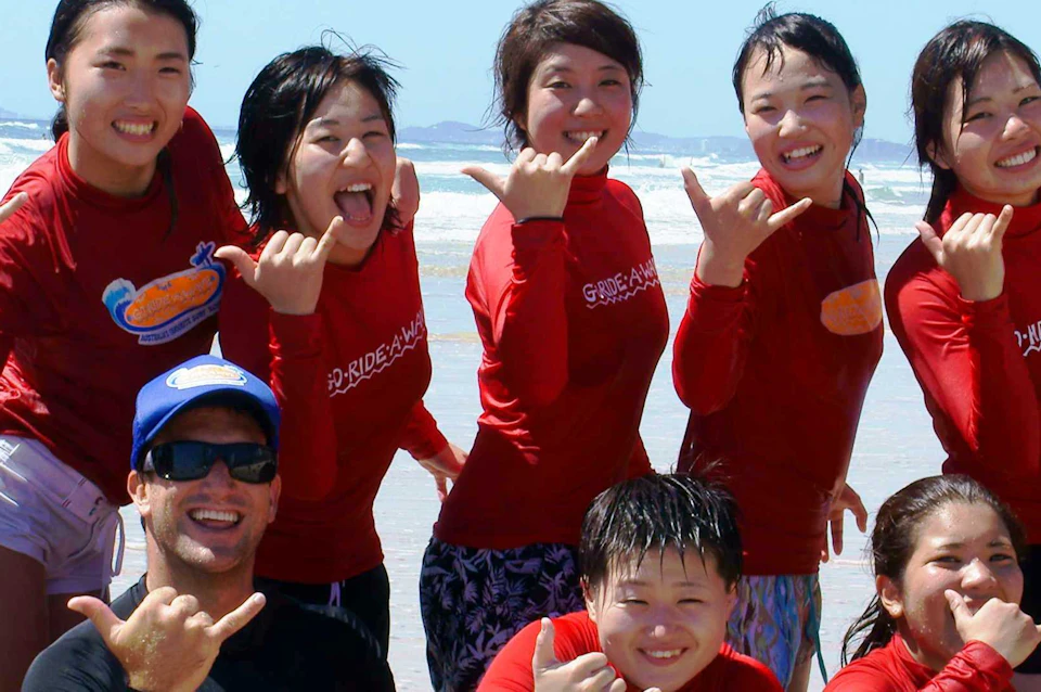 A group of people in red shirts posing with surfboards on a beach, making shaka signs and smiling. Waves are visible in the background.