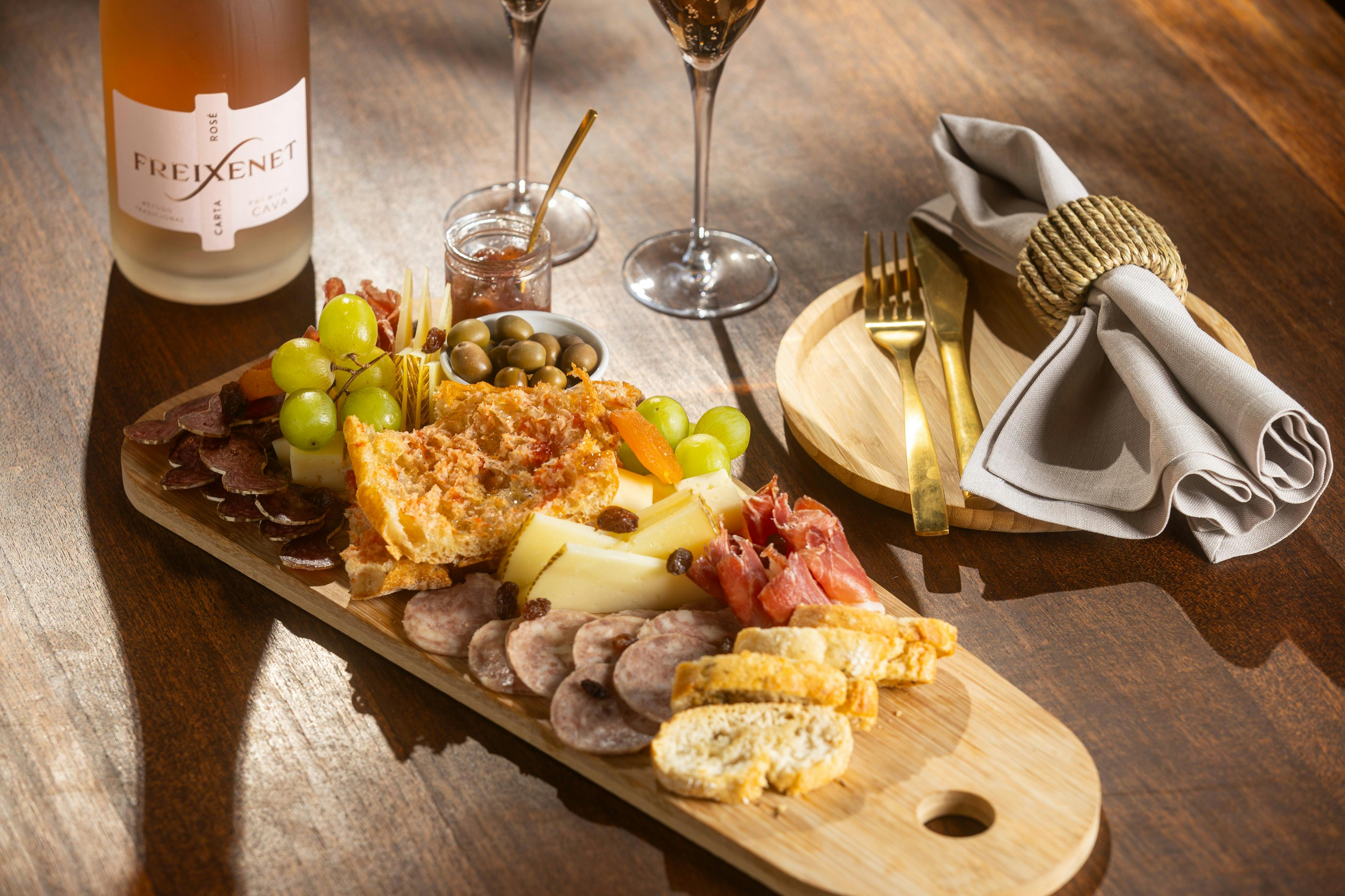 Tabla de embutidos con quesos variados, carnes, uvas, aceitunas, pan y un tarro de mermelada. Cerca, copas de vino, cubiertos y una servilleta.