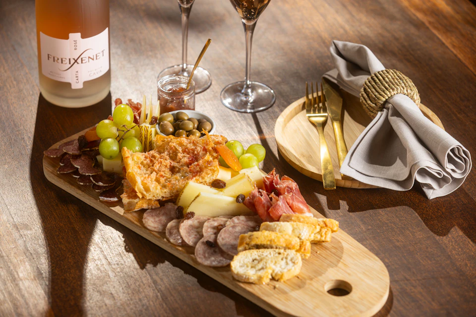Charcuterie board with assorted cheeses, meats, grapes, olives, bread, and a jar of jam. Nearby, wine glasses, cutlery, and a napkin.