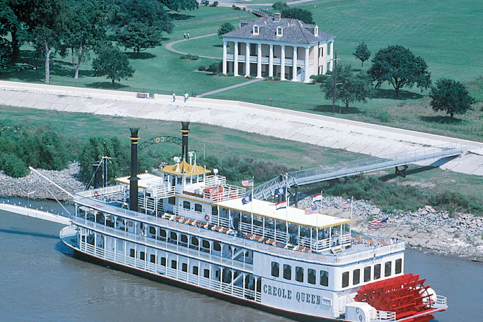 A riverboat named "Creole Queen" floats on a river with a large, white plantation house and green lawn in the background.