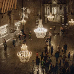 A dimly lit hall with chandeliers, sculptures on stone walls, and a group of people walking and observing the surroundings.