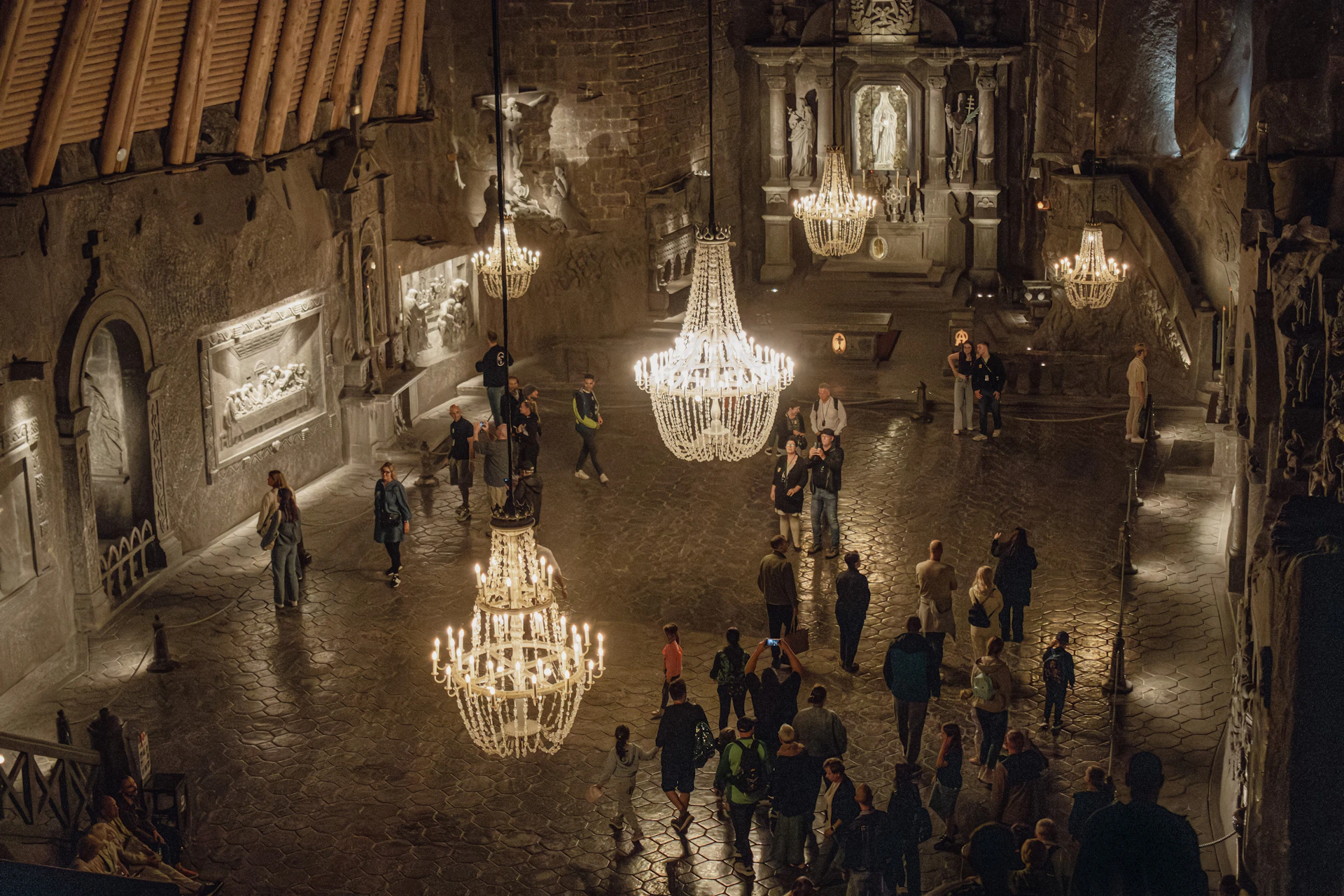 A dimly lit hall with chandeliers, sculptures on stone walls, and a group of people walking and observing the surroundings.