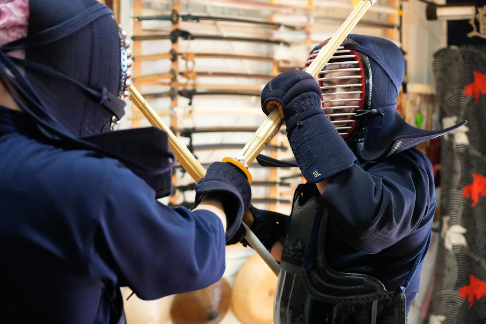 Two individuals in kendo armor practicing with bamboo swords in a dojo, with wooden weapons displayed in the background.