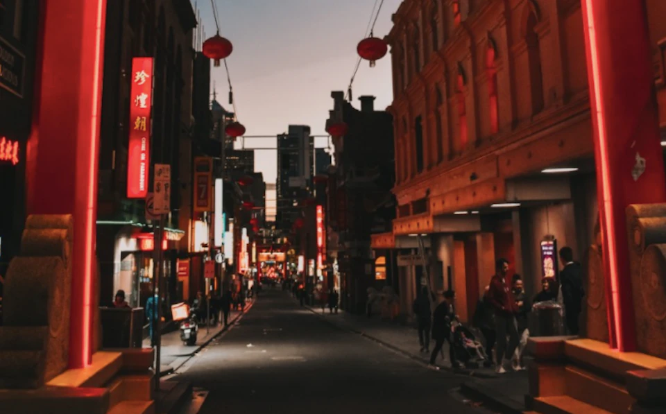 A city street at dusk with lit red lanterns and neon signs, people walking, and buildings on both sides.