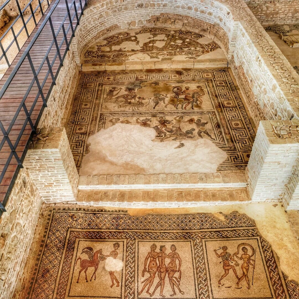 Ancient Roman mosaic depicting scenes with humans and animals, partially damaged, surrounded by brick walls and a metal railing.
