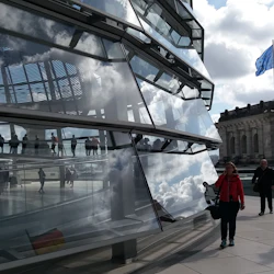 Människor som går inom och utanför en modern glasbyggnad med en reflekterande yta, en gammal stenbyggnad med en blå flagga i närheten.
