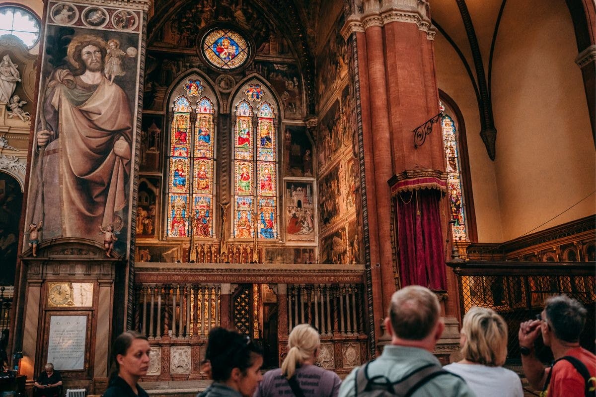 People inside a church with large stained-glass windows, religious paintings, and ornate architectural details in the background.