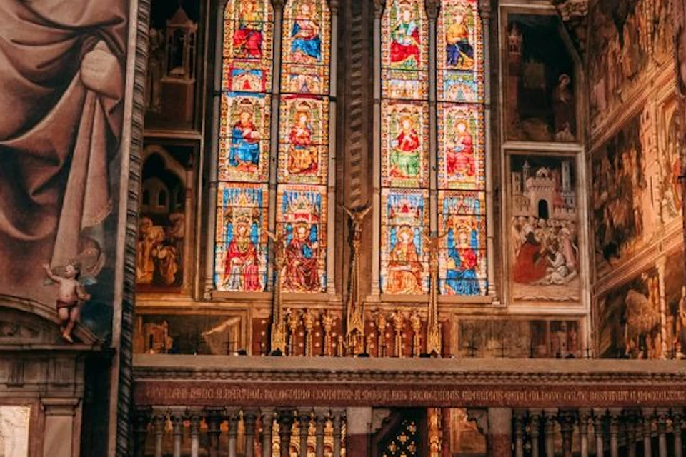 People inside a church with large stained-glass windows, religious paintings, and ornate architectural details in the background.