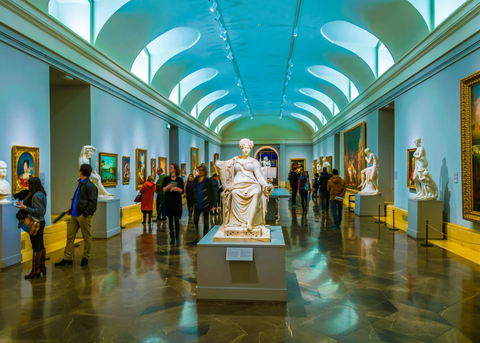 A museum gallery with visitors viewing paintings and sculptures, including a prominent white statue in the centre under arched ceilings.